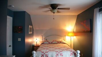 Un dormitorio con cama, ventilador de techo, mesita de noche con lámpara y un cuadro en la pared.