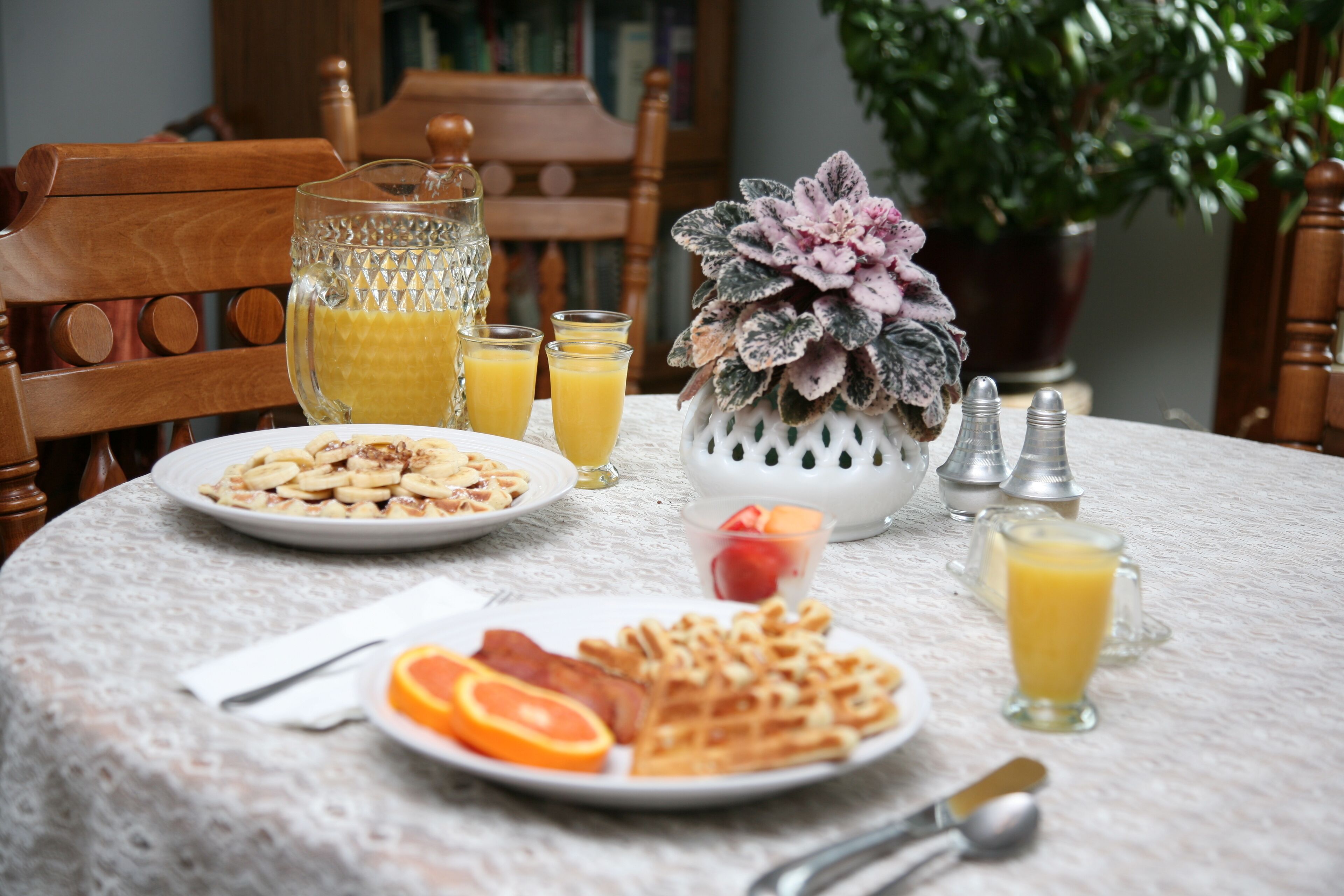 Una mesa de desayuno con platos de gofres, tocino y rodajas de naranja, una jarra de jugo de naranja y un florero con flores.