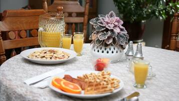 Una mesa de desayuno con platos de gofres, tocino y rodajas de naranja, una jarra de jugo de naranja y un florero con flores.