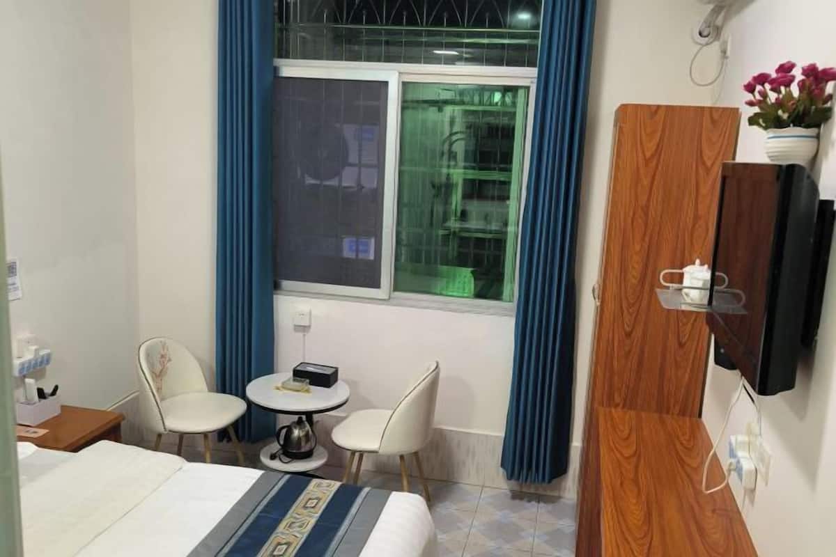 A hotel room with a bed, a small round table, two chairs, a window with blue curtains, a wooden wardrobe, and a wall-mounted TV.