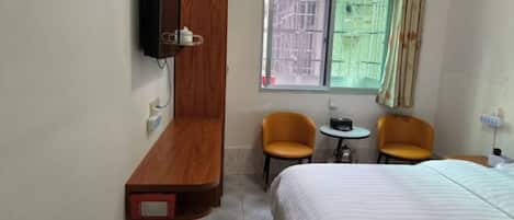 Quarto de hotel com cama, duas cadeiras, uma mesa pequena, TV fixada na parede e janela com vista para o exterior.