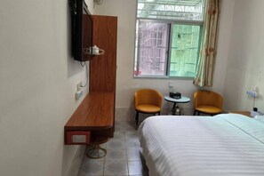 A hotel room with a bed, two chairs, a small table, a TV mounted on the wall, and a window with a view of the outside.