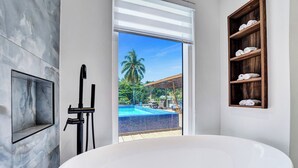 A modern bathroom with a large bathtub, a marble wall, a wooden shelf with towels, and a view of a pool and palm trees. - Spacious 6-bedroom villa - with Outdoor Oasis - Pool in charming Fort Lauderdale (Fort Lauderdale)