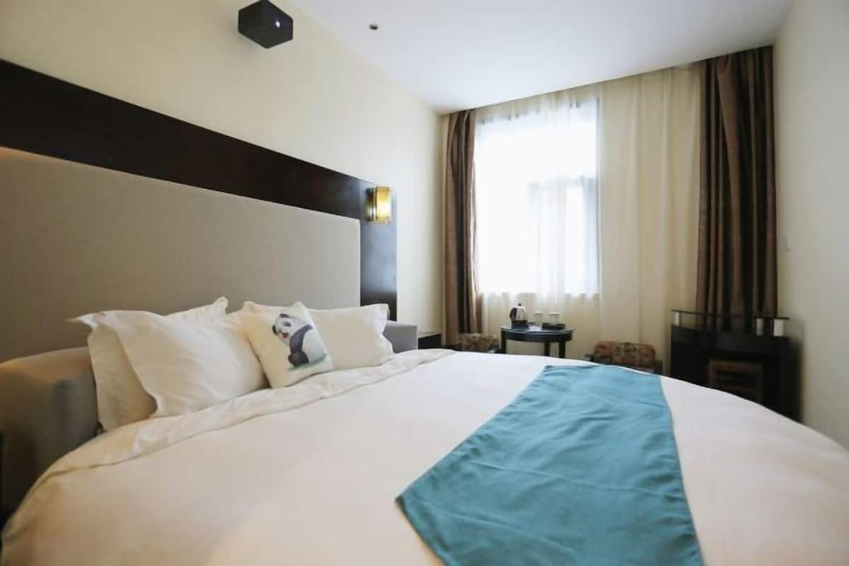 A hotel room with a large bed, a headboard, a small table, a chair, and a window with curtains.