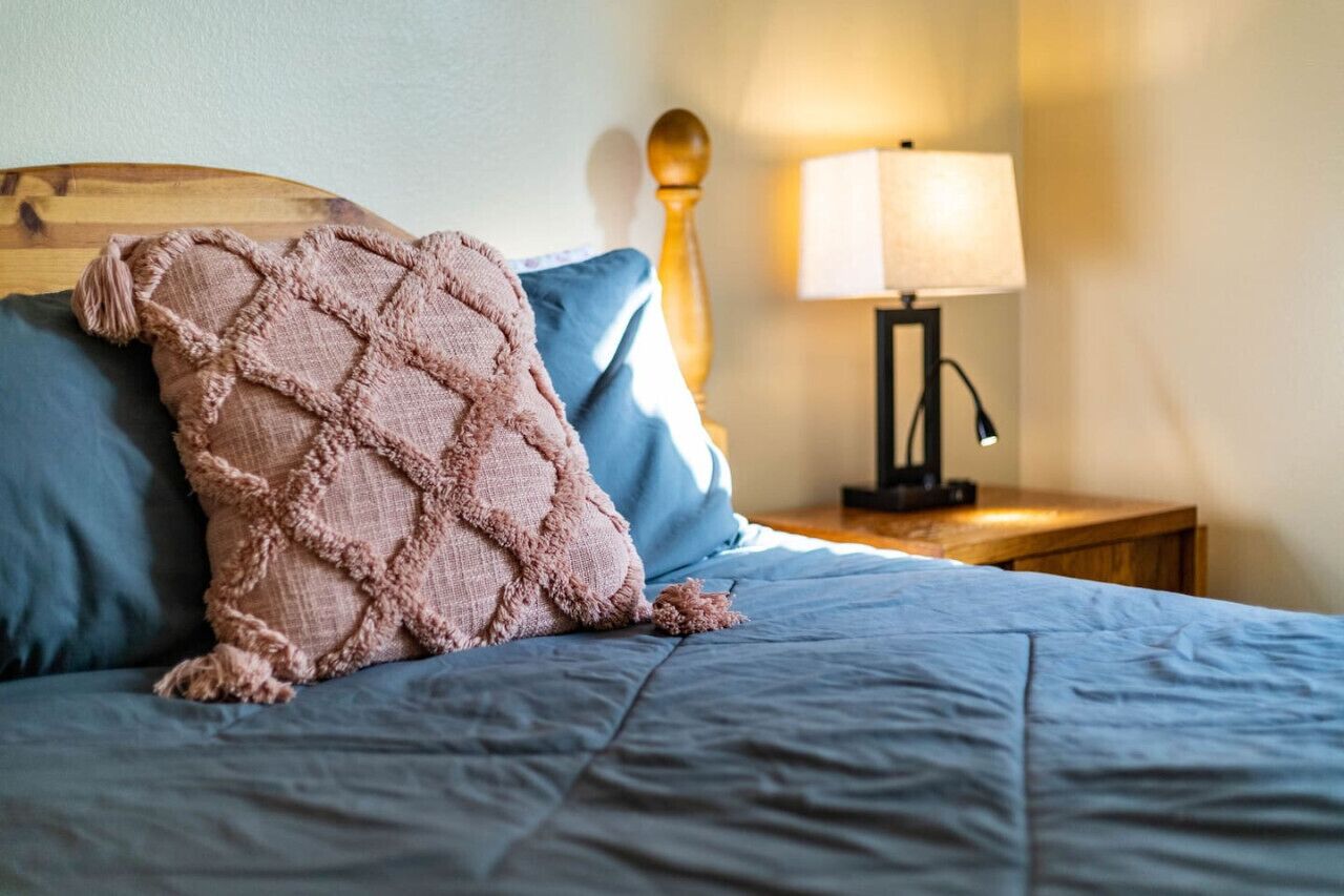 Een net opgemaakt bed met een blauwe plaid en een roze gebreide deken. Er is een houten hoofdbord, een bedlampje en een nachtkastje met een lamp.