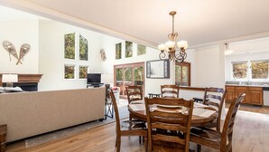 A spacious living room with a large wooden dining table, a sofa, and a fireplace. There are multiple windows providing natural light and views of the outdoors.
