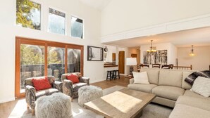 A spacious living area with a large sofa, two armchairs, and a coffee table. There are multiple windows allowing natural light, and a kitchen area is visible in the background.