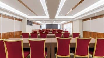 A conference room with rows of red chairs and tables, wooden paneling on the walls, and a flat-screen TV mounted on the wall.