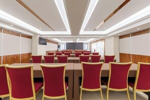 A conference room with rows of red chairs and tables, wooden paneling on the walls, and a flat-screen TV mounted on the wall.