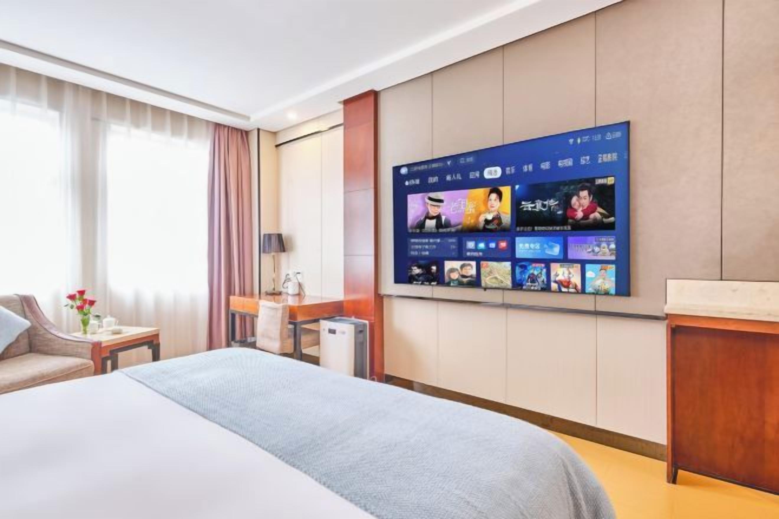 A hotel room with a large flat-screen TV mounted on the wall displaying various content. There is a bed with a grey blanket, a bedside table with a lamp, and a small table with a vase of flowers.