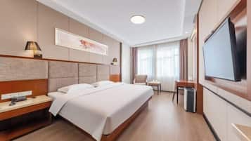 A hotel room with a bed, bedside table, lamp, TV, and a window with curtains.