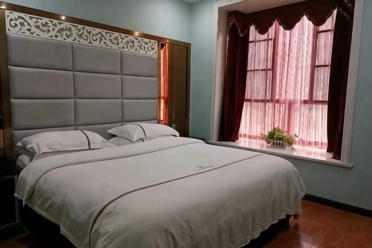 A bedroom with a large bed, a headboard with a decorative panel, a window with curtains, and a small plant on the windowsill.