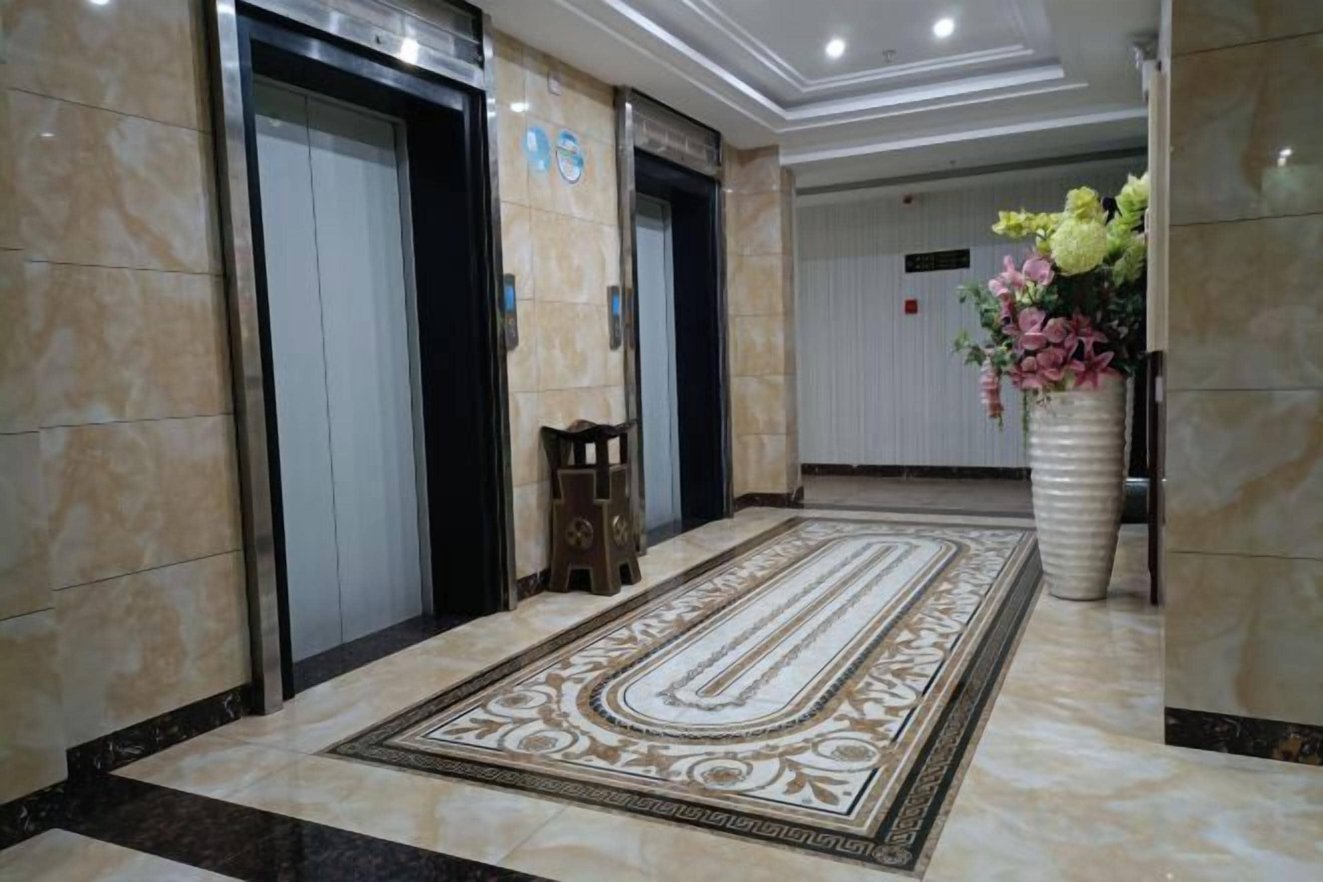 A hotel lobby with marble flooring, two elevators, a decorative rug, and a vase of flowers.
