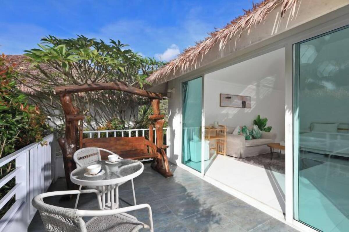 Ein Balkon mit einer Holz-Pergola, weißen Korbmöbeln und einer Glastür zu einem Wohnbereich mit einer Couch und einem Couchtisch.