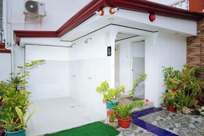 Entrada de hotel con fachada blanca, tejado rojo y suelo de baldosas. Hay plantas en macetas y un felpudo verde en la entrada.