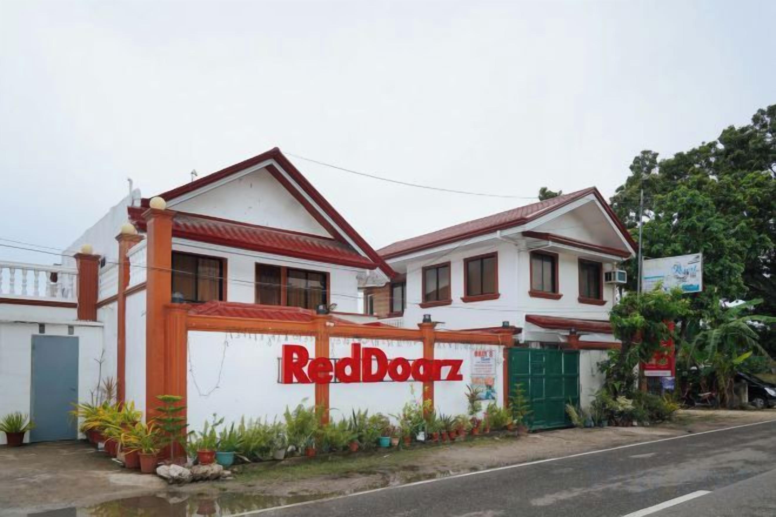 A two-story building with a red and white facade, a prominent RedDoorz sign, and a gated entrance.