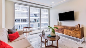 A modern living room with a sofa, a coffee table, a TV mounted on the wall, and a large window with a view of buildings. - Quinta San Miguel Canal Pool Steps Beach (Nuevo Nayarit)