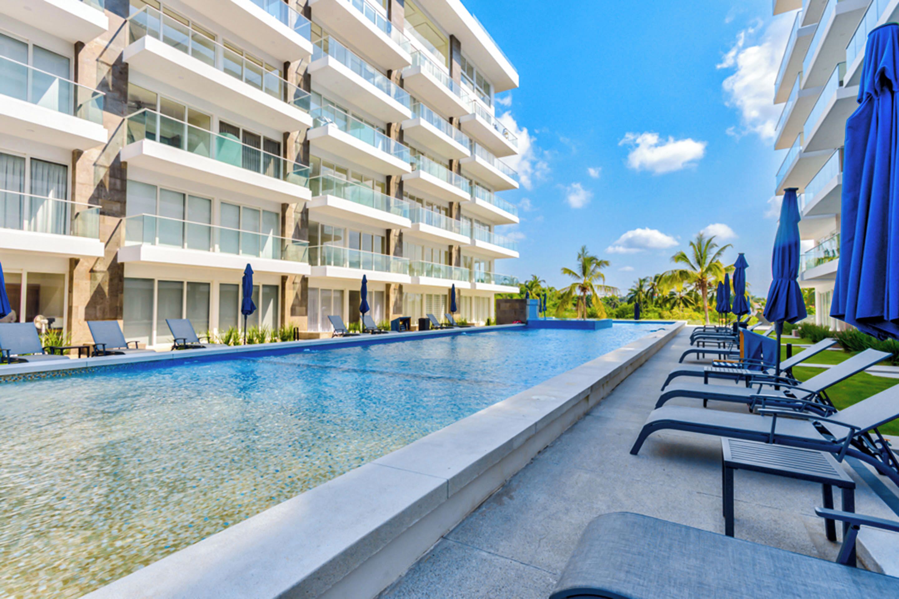 A modern hotel with a swimming pool, sun loungers, and blue umbrellas.