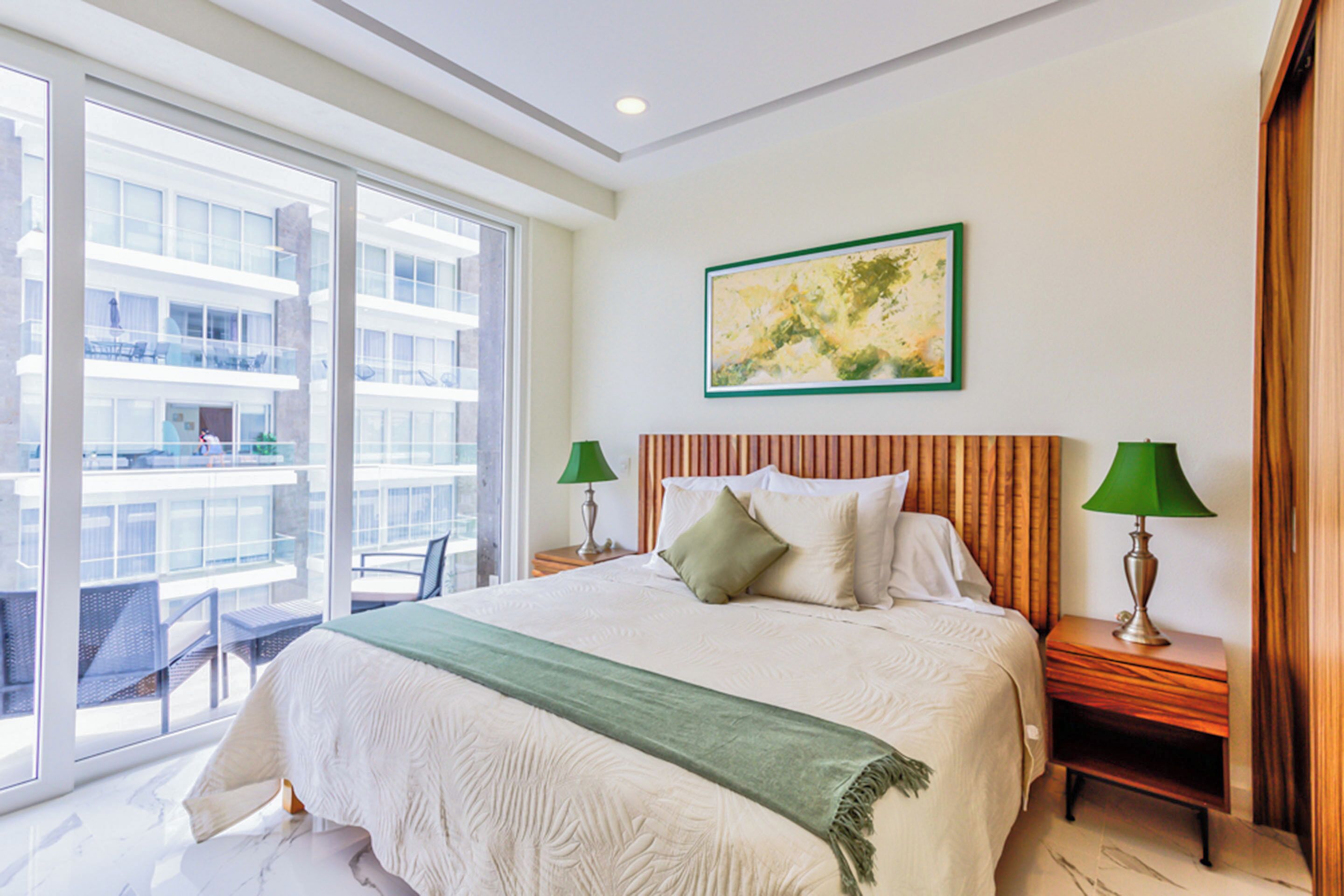 A hotel room with a large bed, two bedside tables with lamps, a green throw blanket, and a large window offering a view of a building.