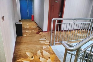 Un pasillo con alfombra, barandilla de metal, una puerta roja y un bote de basura.