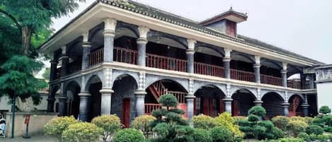 A traditional building with multiple balconies and a tiled roof, surrounded by a garden with various plants.