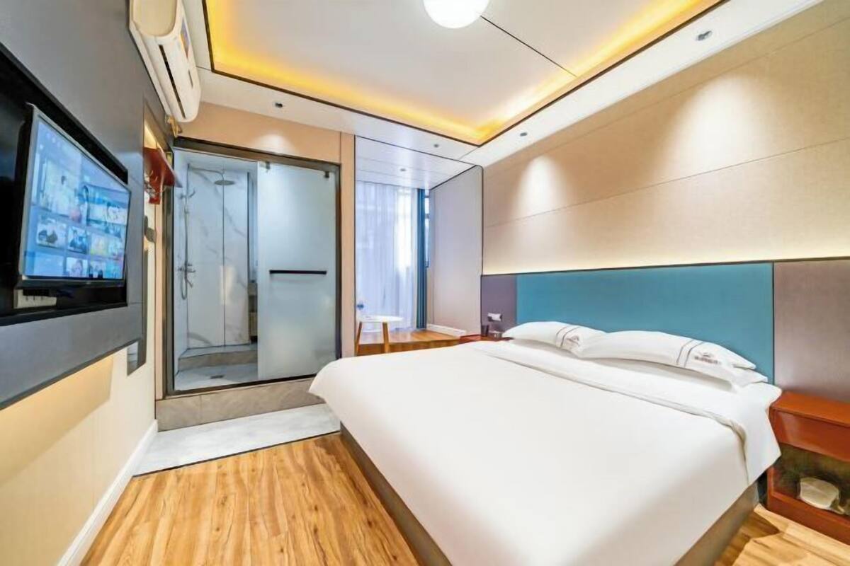 A modern hotel room with a large bed, a television, a glass shower enclosure, and a wooden floor.