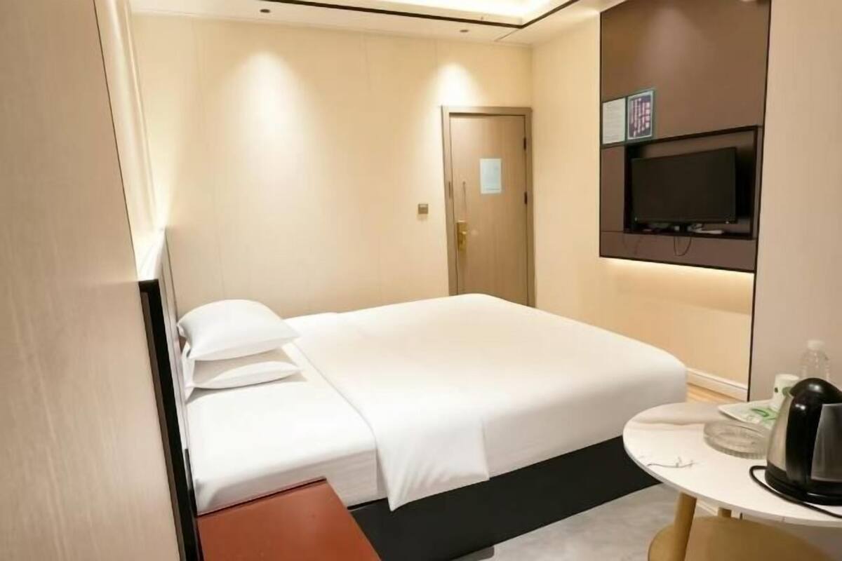 A hotel room with a bed, a television, a small table with a kettle, and a door.