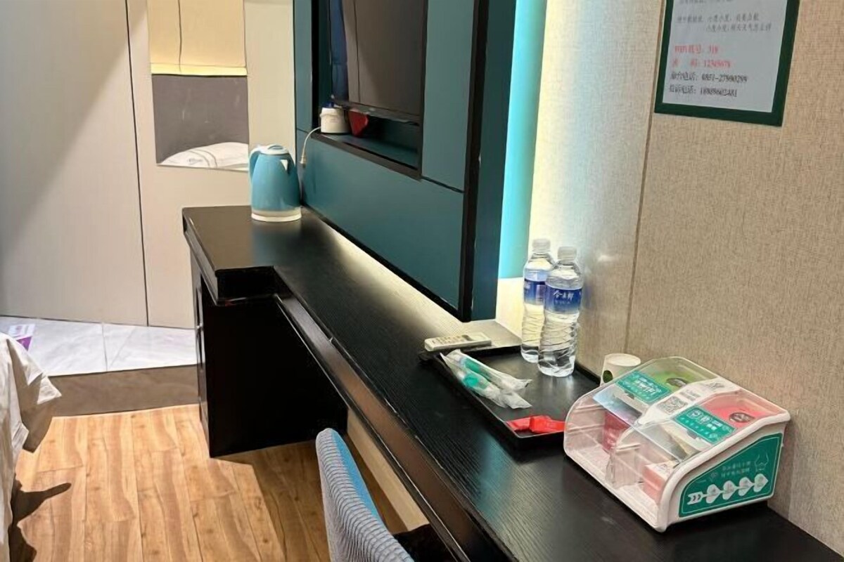 A hotel room with a wooden floor, a black desk, a blue kettle, a water bottle, a tissue box, and a tea set on the counter.