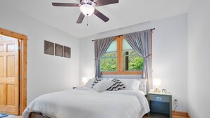 A bedroom with a bed, nightstands, a ceiling fan, a window with curtains, and a wooden door.