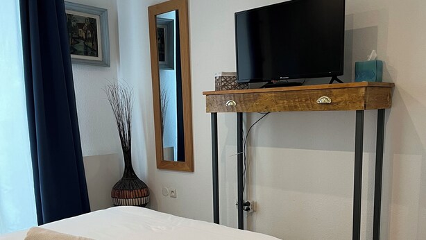 Un lit bien fait, avec une serviette enroulée, une télévision posée sur une table en bois et un miroir accroché au mur.