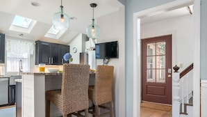A kitchen with dark cabinets, a breakfast bar with wicker chairs, a TV mounted on the wall, and a wooden staircase leading to a door. - Onset Oasis_2 min walk to beach, Sleeps 14, HUGE yard w/ lawn games & 2 grills! (Wareham)