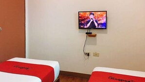 Een hotelkamer met twee bedden, elk met een rode hoofdbord en een witte matrasbeschermer. Een televisie is aan de muur bevestigd, waarop iemand zingt.