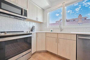 Een moderne keuken met een inbouwoven, magnetron en vaatwasser, een raam met uitzicht op een huis en lichtgekleurde keukenkastjes.