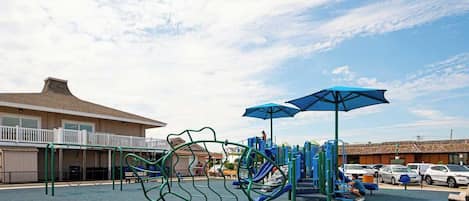 A playground with climbing structures and slides, a covered seating area, and a building with a balcony in the background.