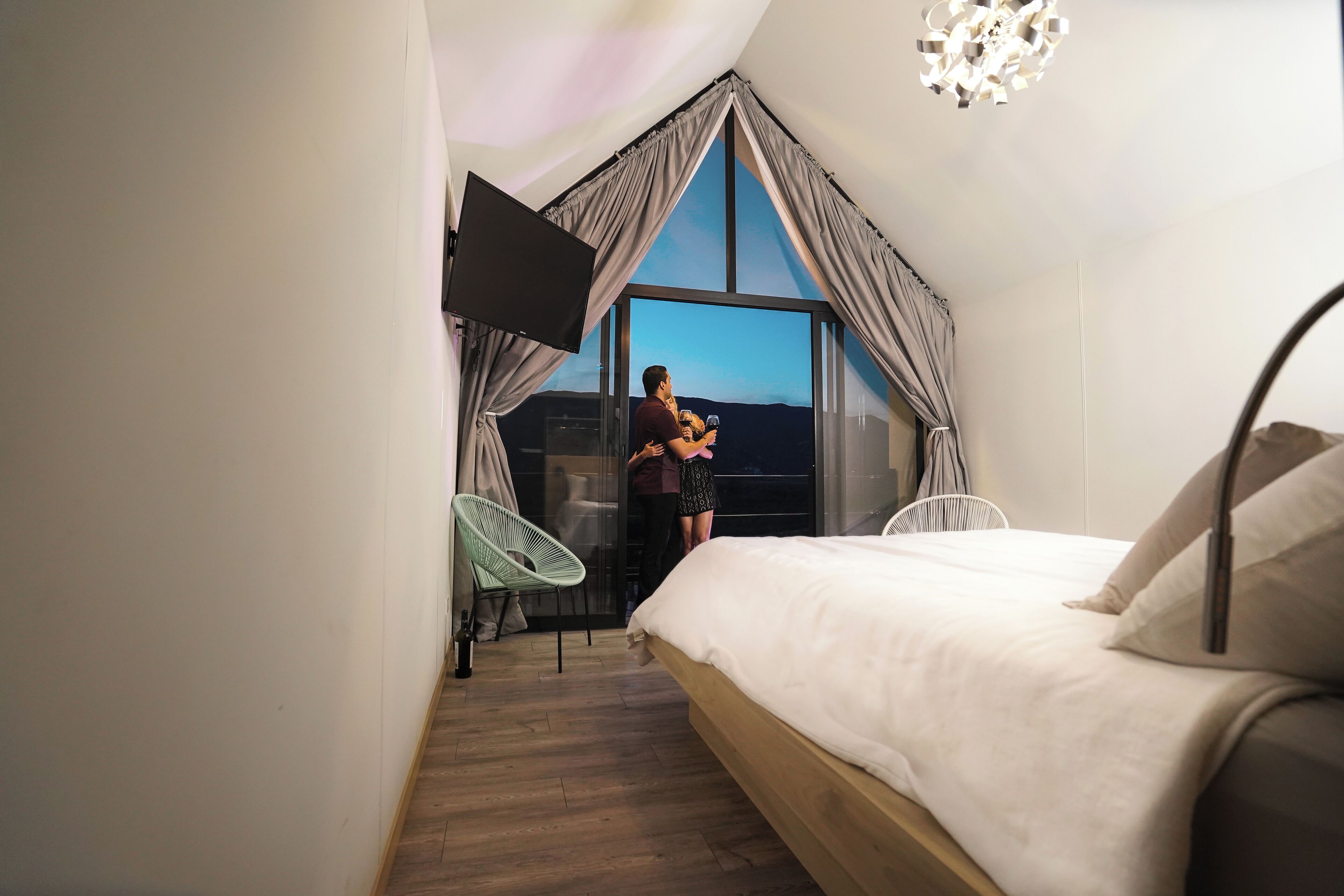 Un dormitorio con un ventanal grande, una cama, un sillón y un televisor montado en la pared.