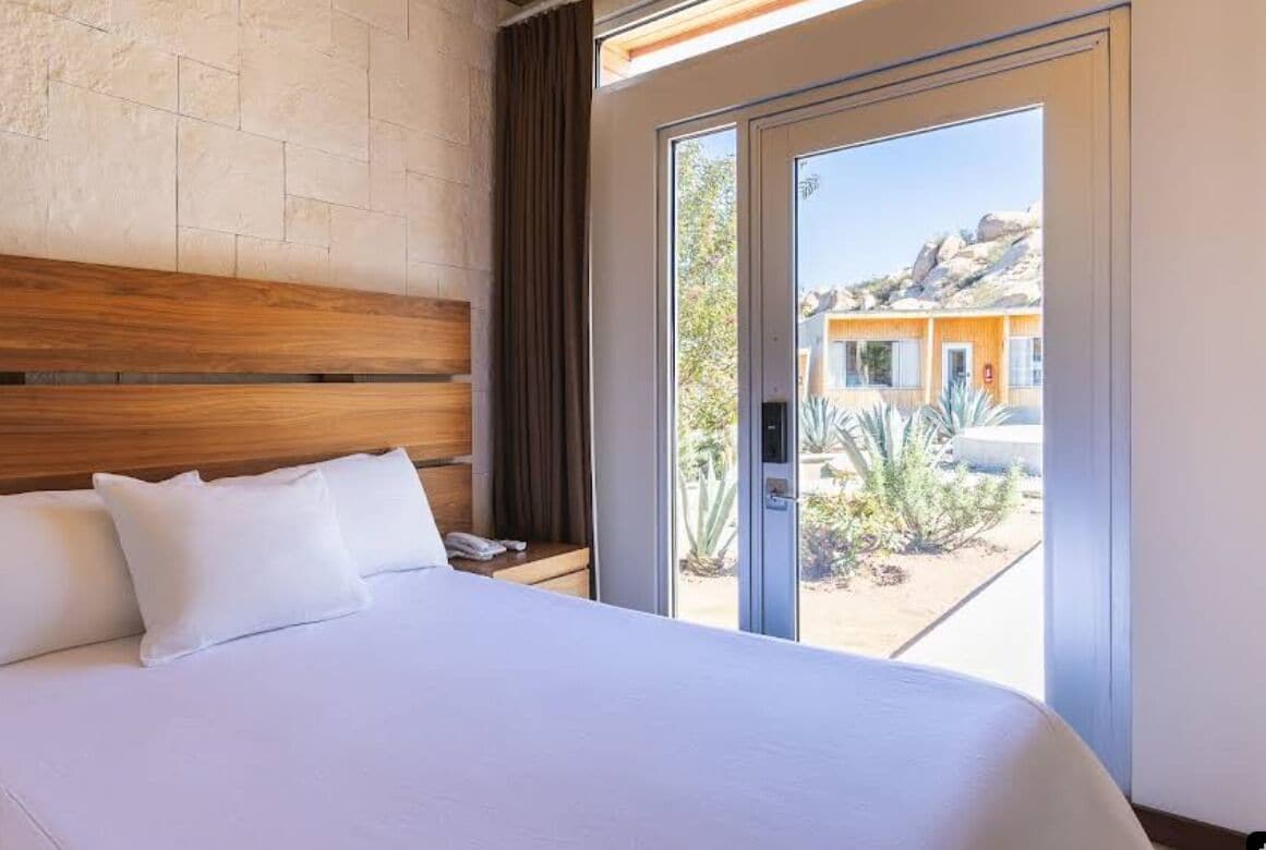 Habitación de hotel con una cama grande, cabecera de madera y puerta corrediza de vidrio que da a un espacio exterior con vistas a un edificio y vegetación desértica.
