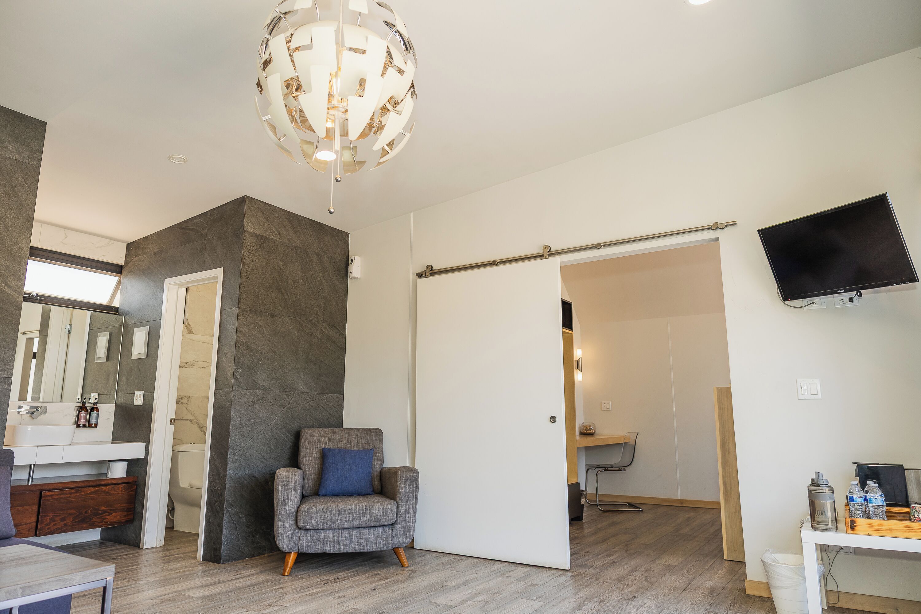 A modern bathroom with a large mirror, a grey armchair, and a sliding door leading to another room.