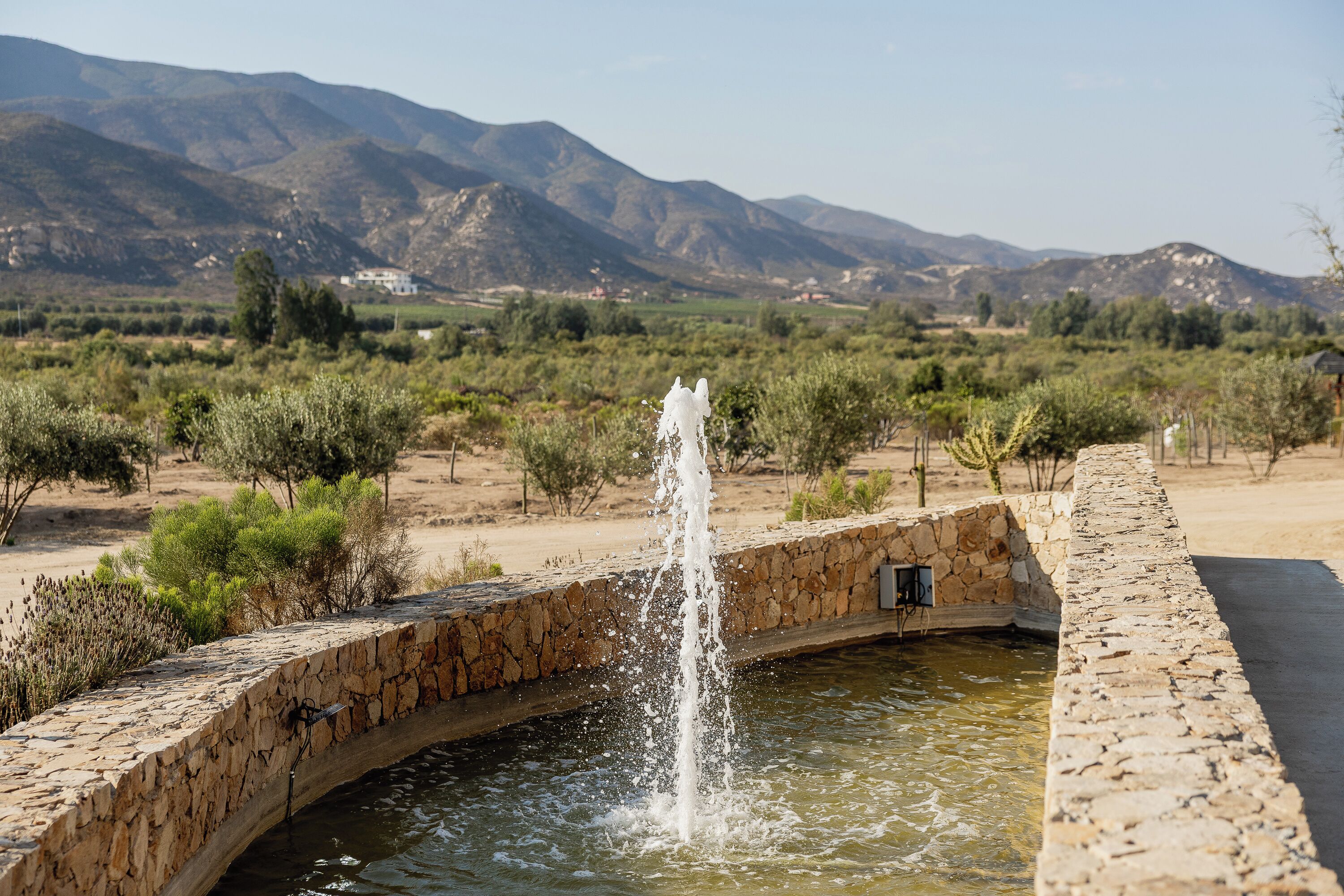 A stone bridge over a body of water with a fountain in the center, surrounded by a desert landscape with mountains in the background.