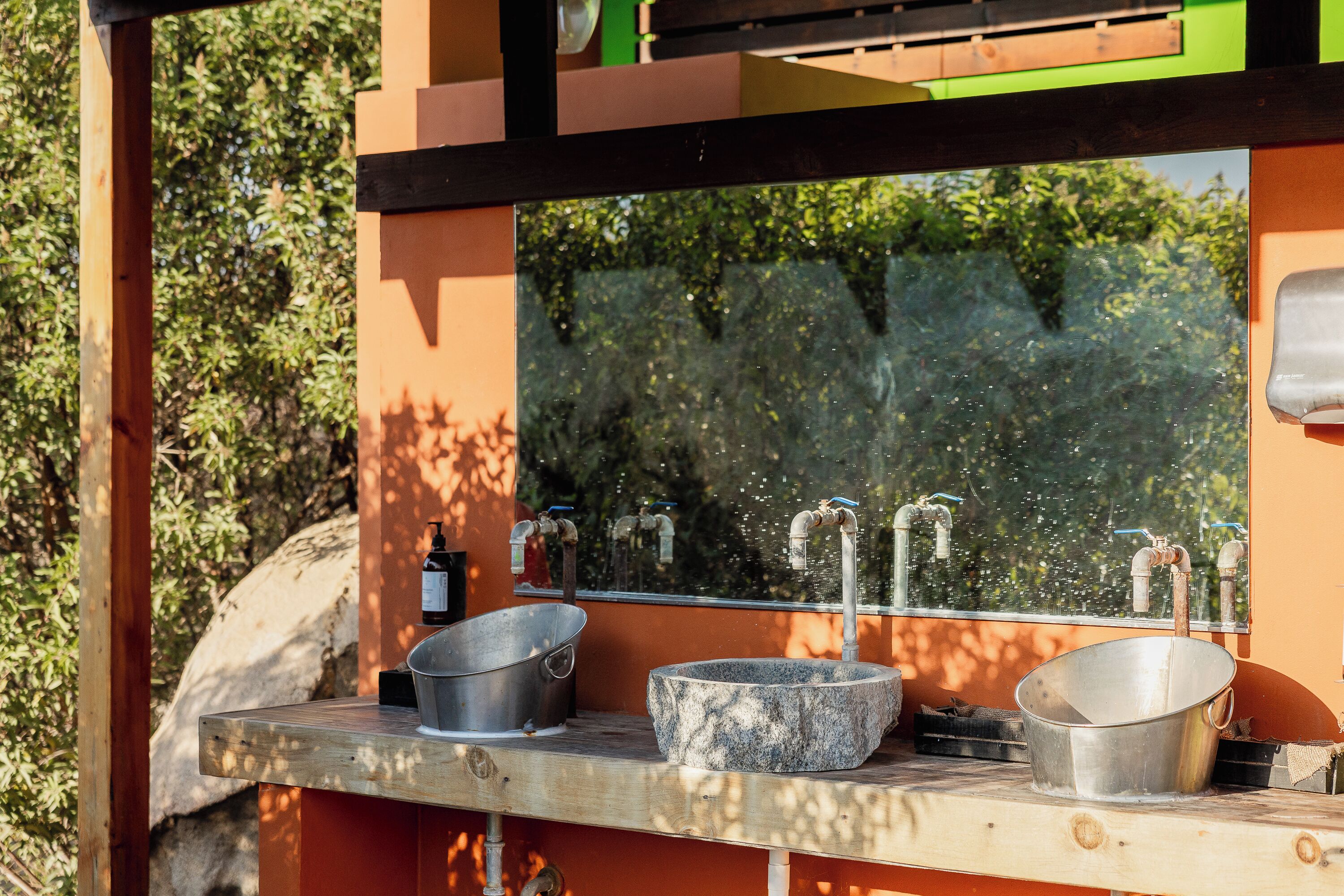 A rustic outdoor sink area with a stone basin, a metal basin, and a bottle of soap.