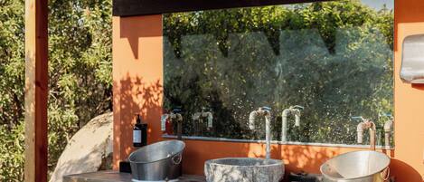 A rustic outdoor sink area with a stone basin, a metal basin, and a bottle of soap.