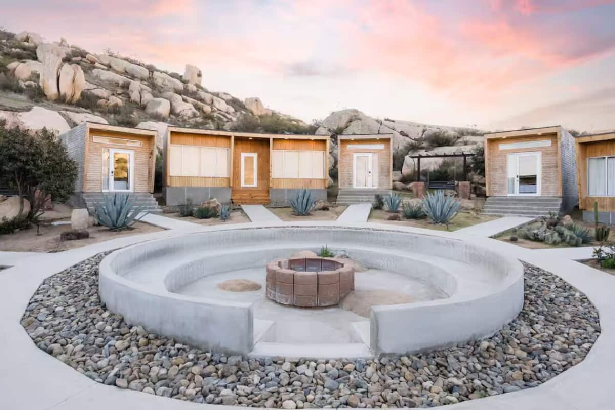 A modern outdoor area with a circular stone fire pit surrounded by a concrete ring, and four wooden cabins with large windows and doors.