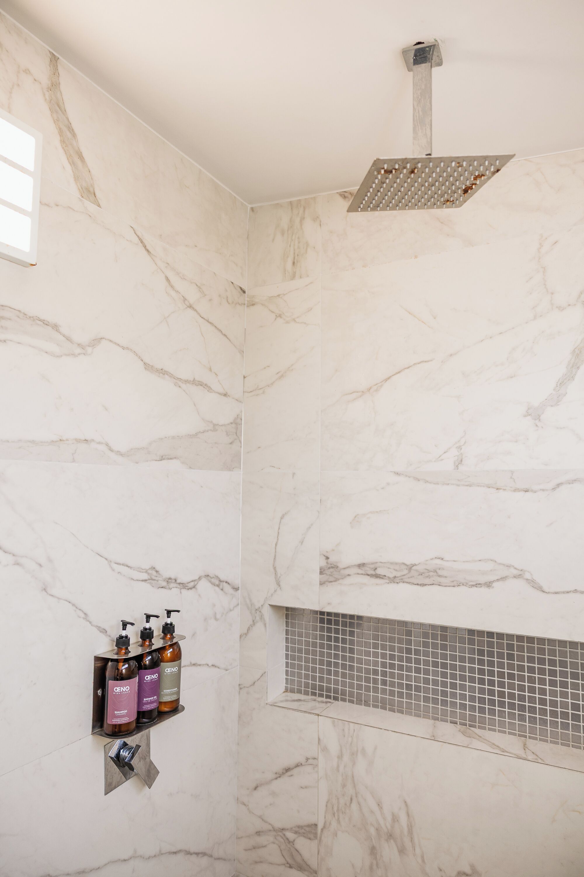 Un baño con paredes de mármol, una ducha y artículos de aseo en un estante.
