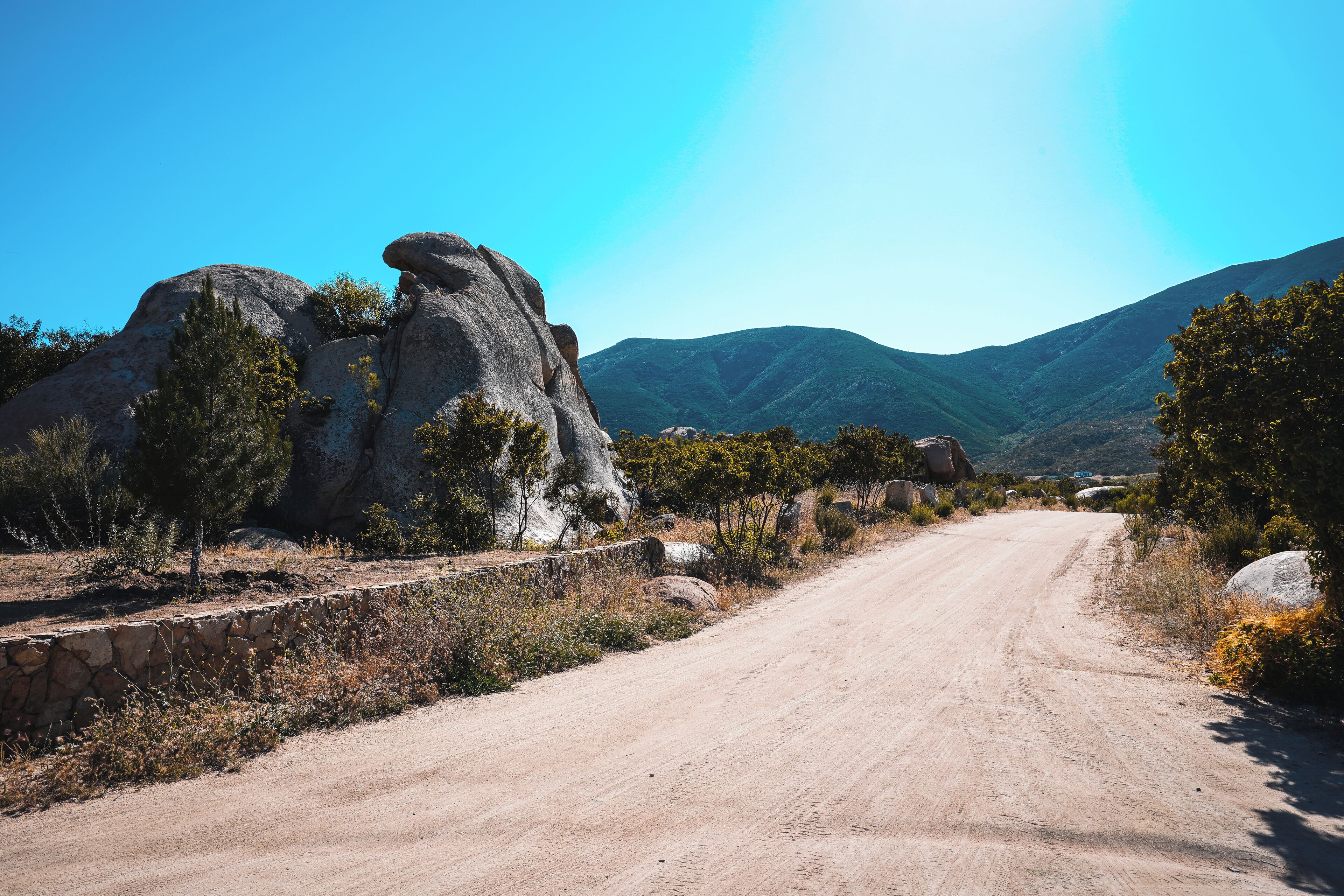 A dirt road flanked by large boulders and sparse vegetation, leading towards a mountainous landscape under a clear blue sky.
