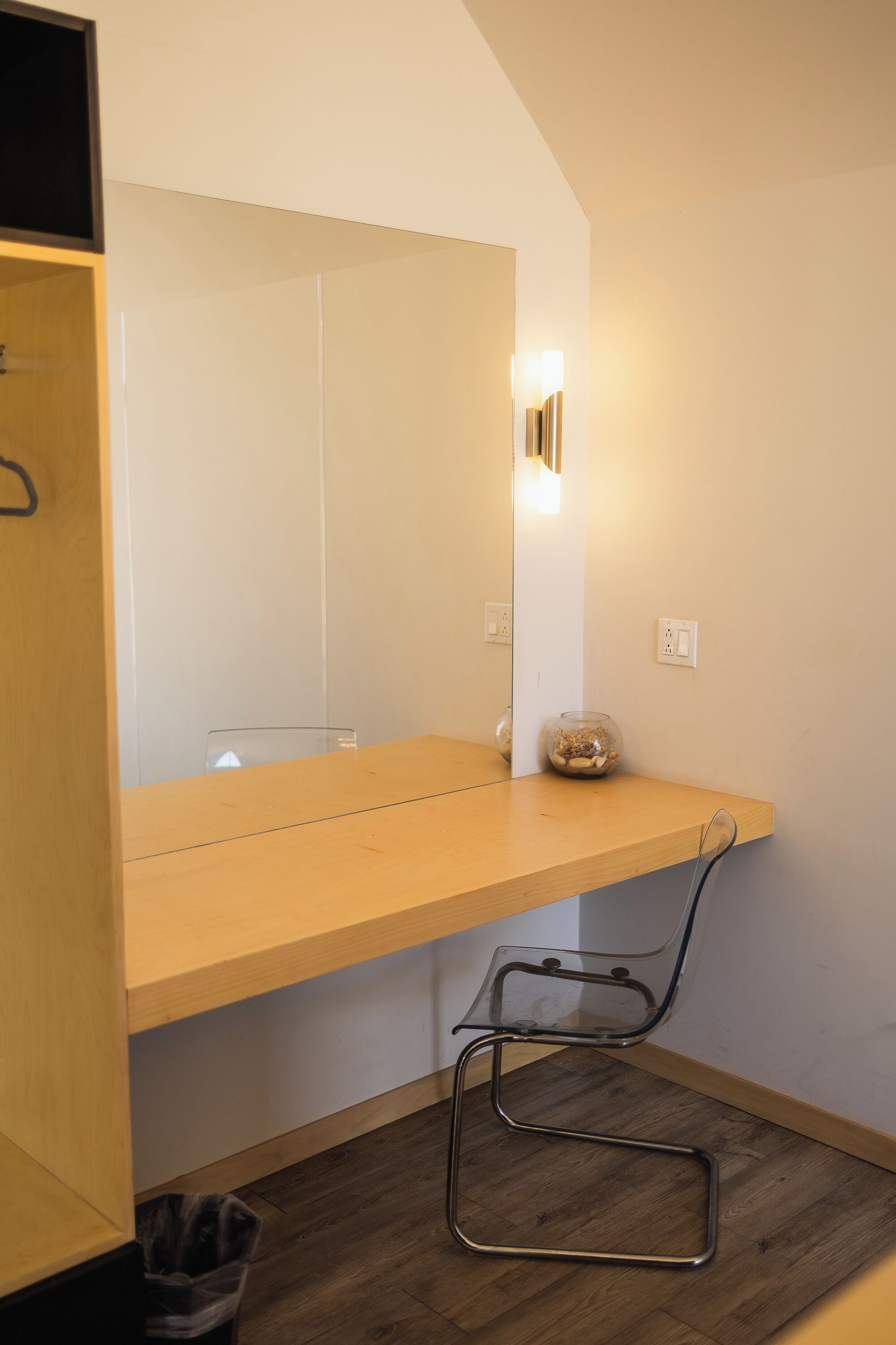 A small, modern bathroom with a wooden vanity, a glass chair, and a mirror with a light fixture.