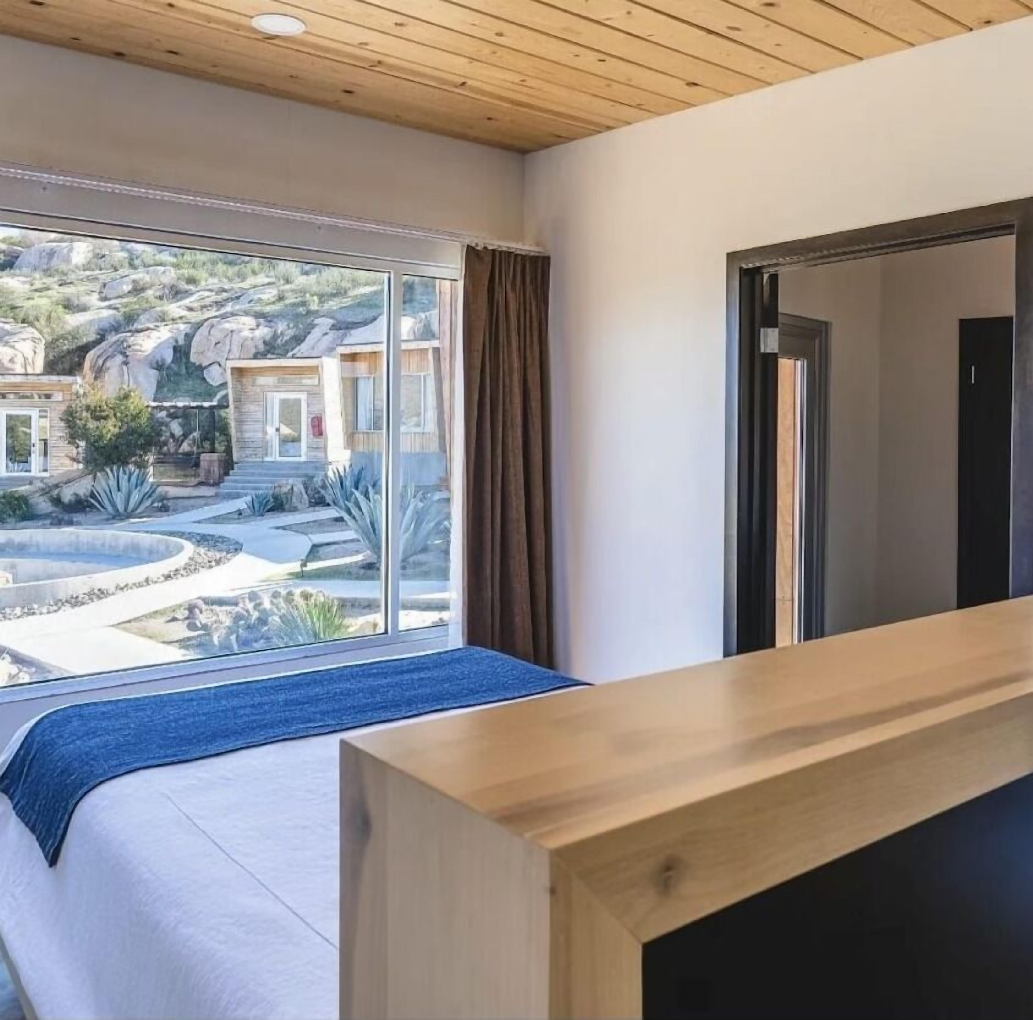 Une chambre moderne dotée d’une grande fenêtre, d’une tête de lit en bois et donnant sur un espace extérieur aménagé.