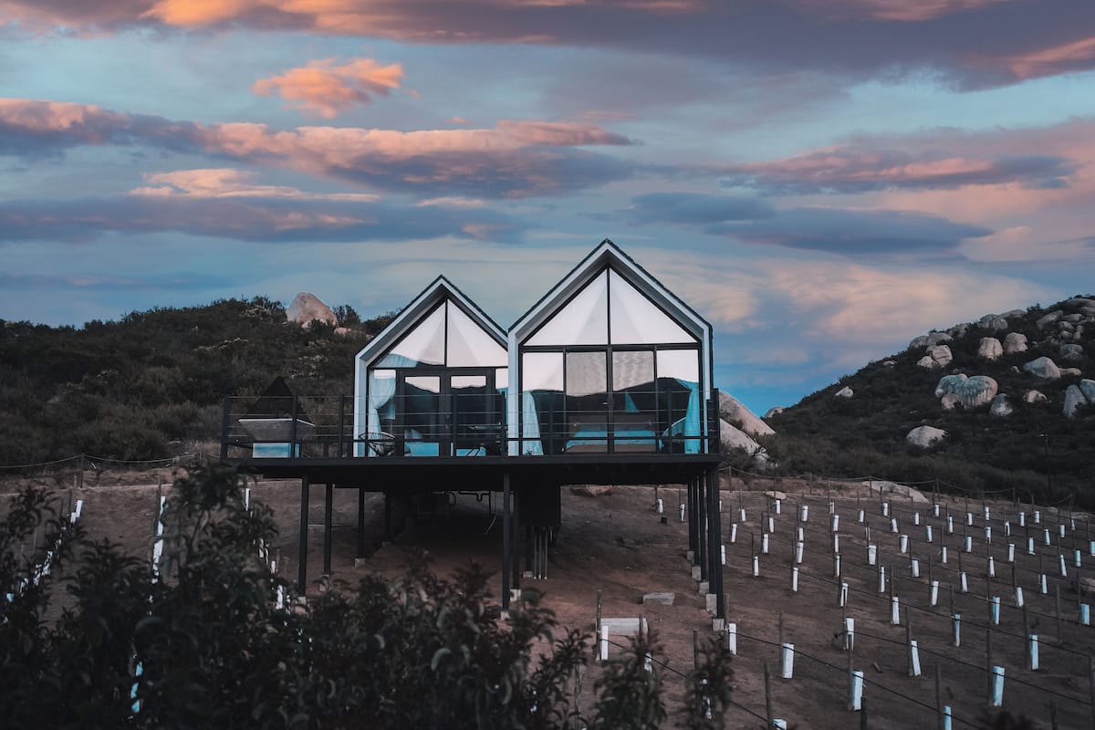 A modern, two-story building with a glass facade and a balcony, situated on a hill with a rocky landscape in the background.