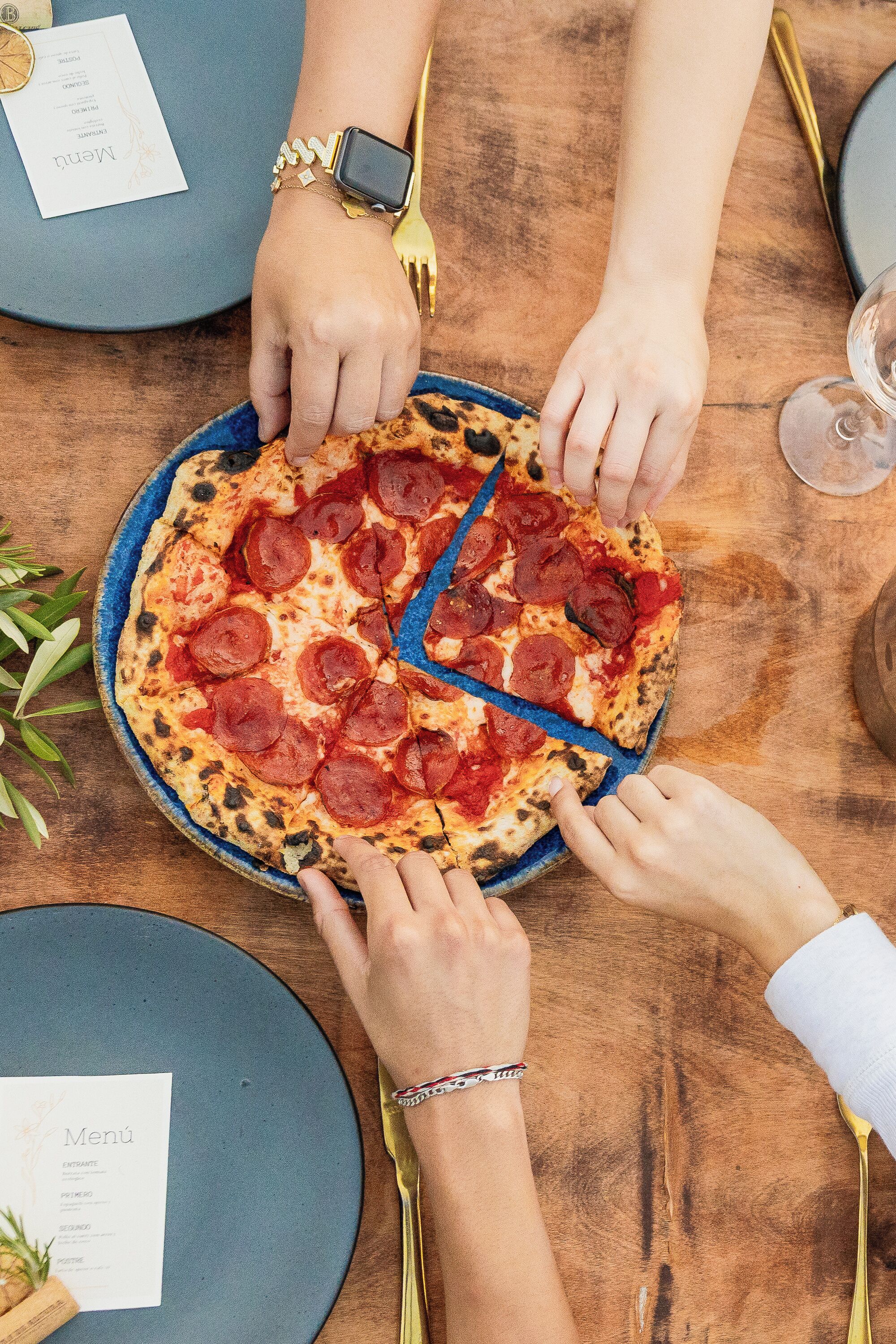 A pizza being shared among four people at a table with a menu and a wine glass.