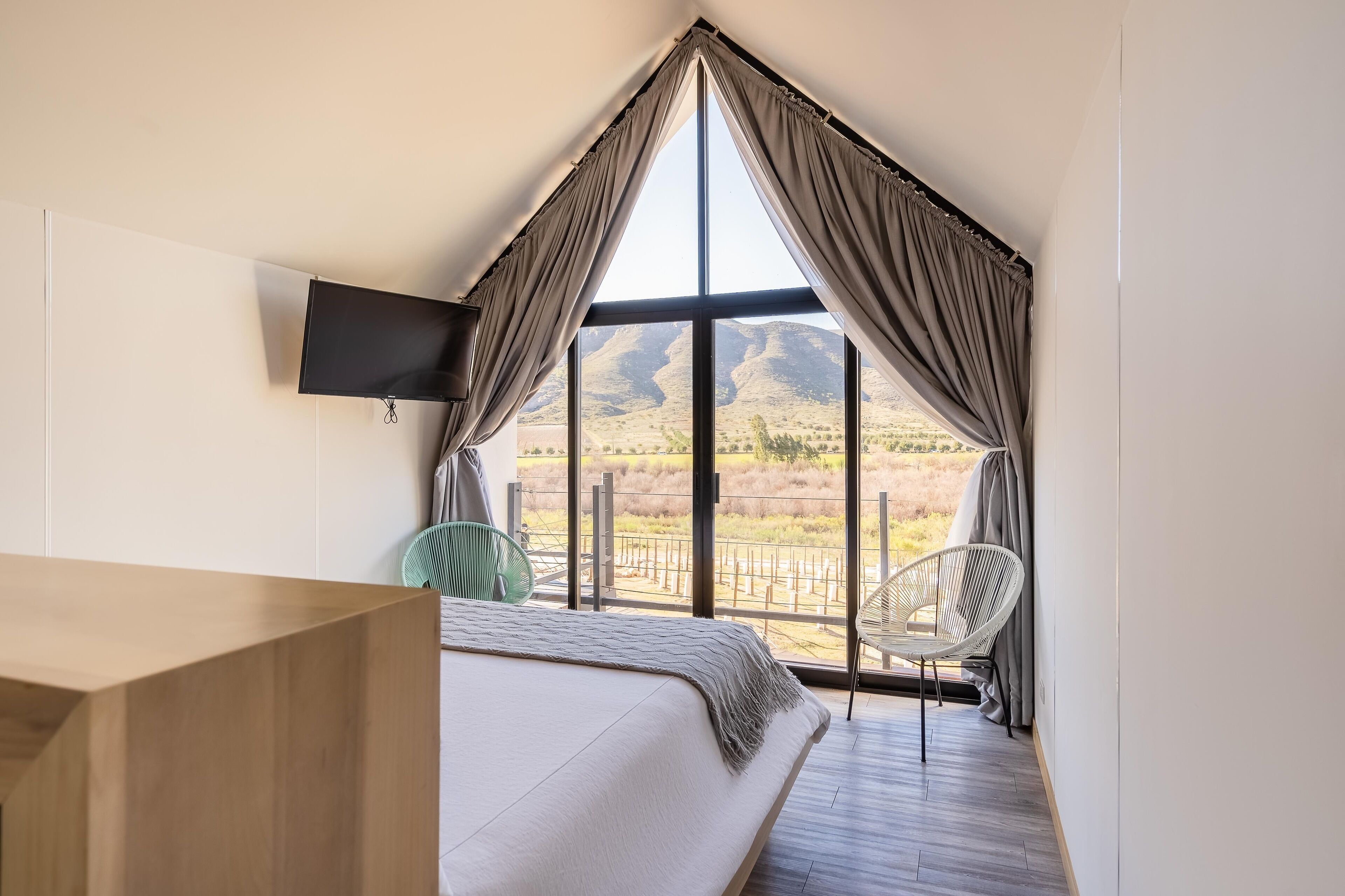 Ein Schlafzimmer mit großem Fenster, einem Bett, einem an der Wand montierten Fernseher und einem Sessel.