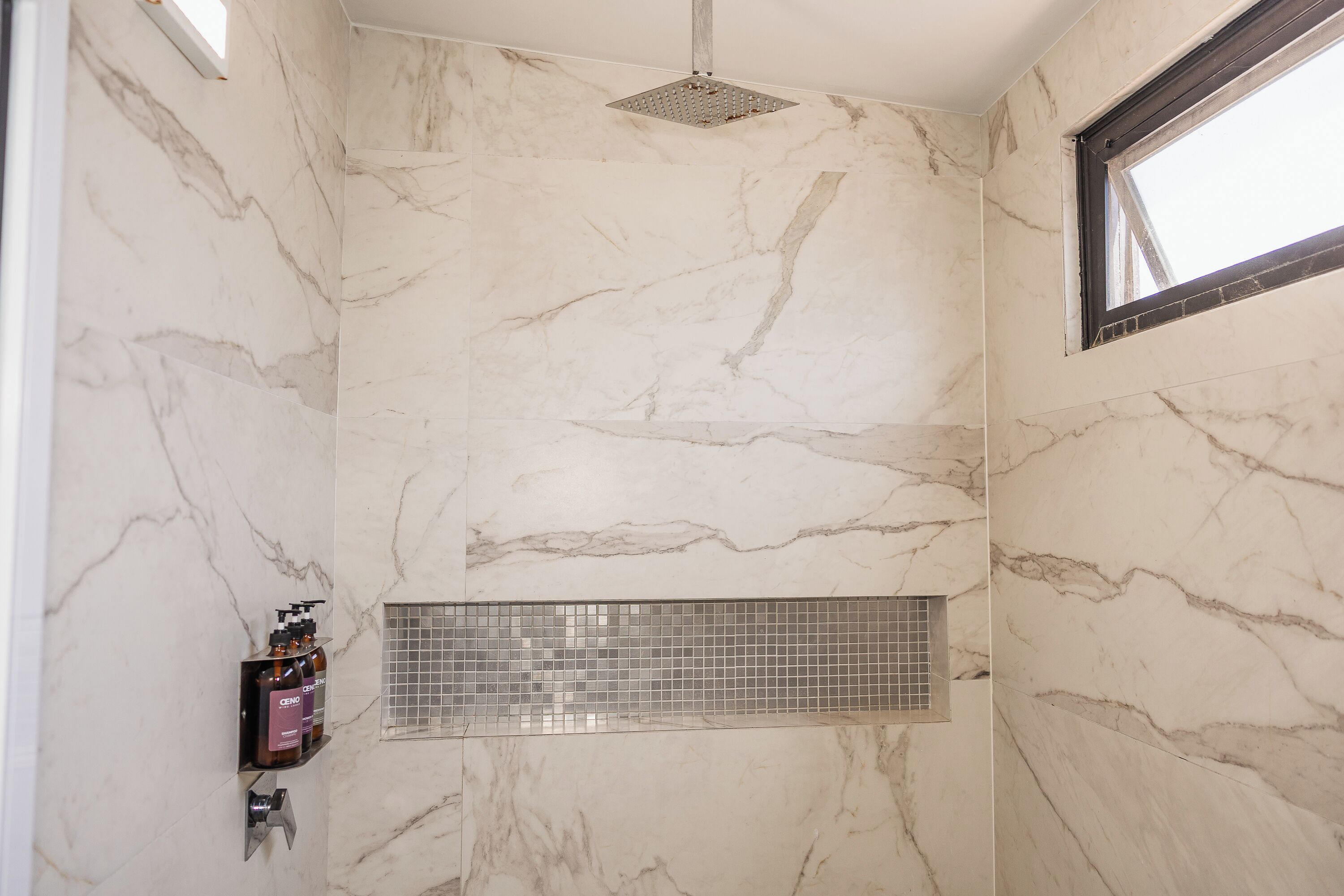 A bathroom with marble walls, a shower with a rain showerhead, a window, and a shelf with toiletries.
