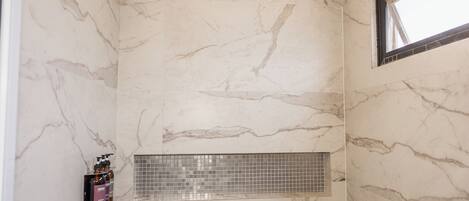 A bathroom with marble walls, a shower with a rain showerhead, a window, and a shelf with toiletries.